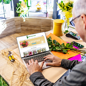 Man typing on laptop in bright flower shop, browsing Michelle's Flowers Limited website with bouquets and floral supplies