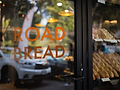 A glass bakery door features bold orange lettering spelling "ROAD BREAD." Through the reflection, a parked white car and greenery are visible. Inside, shelves lined with neatly arranged baguettes add to the inviting, artisanal feel of the shop.