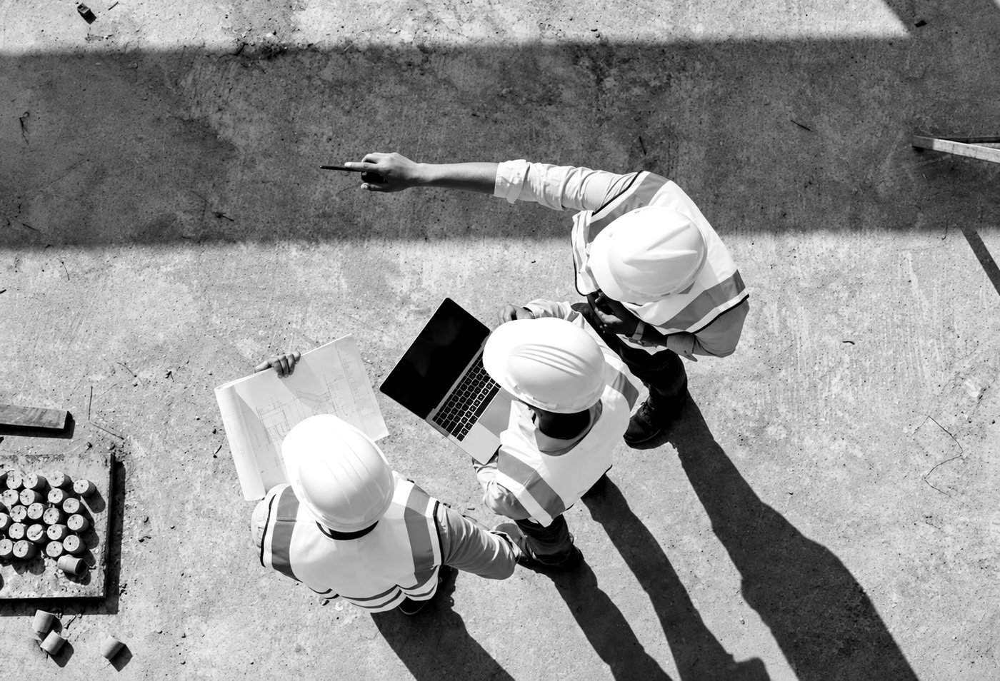 Overhead black and white view of three site workers in hard hats and hi-vis reviewing printed plans and a laptop together