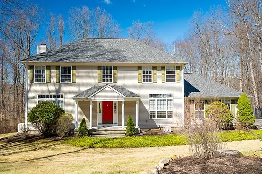 Spacious beige house with red door, green shutters, grey roof, and wooded background..webp