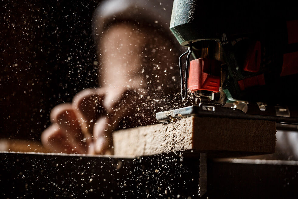 A imagem mostra uma pessoa usando uma serra tico-tico elétrica laranja para cortar um pedaço de madeira, usando luvas de proteção laranja e cinza e roupa de trabalho azul.