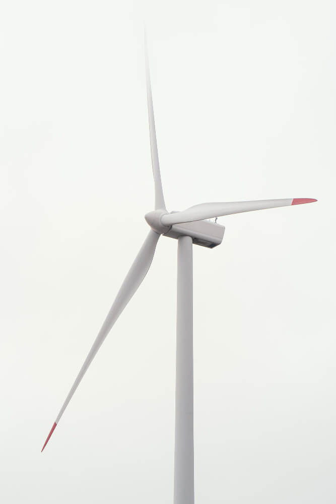 A imagem mostra uma turbina eólica vista de baixo para cima, destacando sua estrutura alta e esguia. O mastro é branco e sustenta um conjunto de três pás longas, levemente curvas, com pontas destacadas em vermelho, detalhe comum para visibilidade aérea.