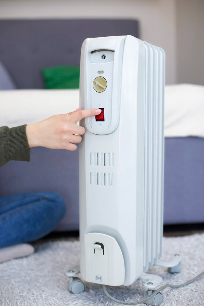 A imagem mostra uma pessoa ligando um aquecedor elétrico portátil. O aparelho é branco, tem rodinhas na base e um botão vermelho de ligar/desligar na parte frontal. Ele está em um ambiente interno, com carpete e uma cama ao fundo.