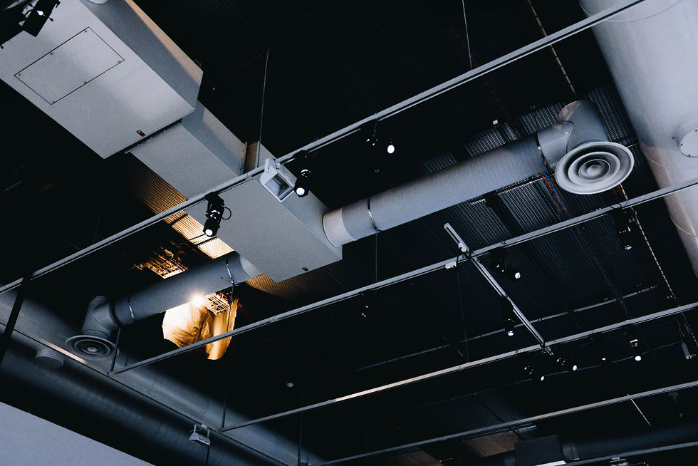 Teto industrial com dutos e ventilação cinza sobre fundo preto, luzes pendentes e brilho quente em uma abertura no alto.