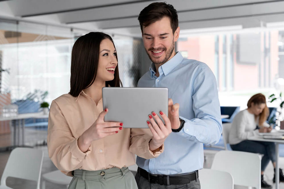 um homem e uma mulher em um ambiente corporativo moderno e iluminado, segurando um tablet e interagindo de forma animada. A mulher, vestindo uma blusa bege e calça verde, sorri enquanto mostra algo na tela do dispositivo para o homem, que usa uma camisa azul clara e um relógio no pulso. Ambos parecem engajados e satisfeitos com o que estão vendo.