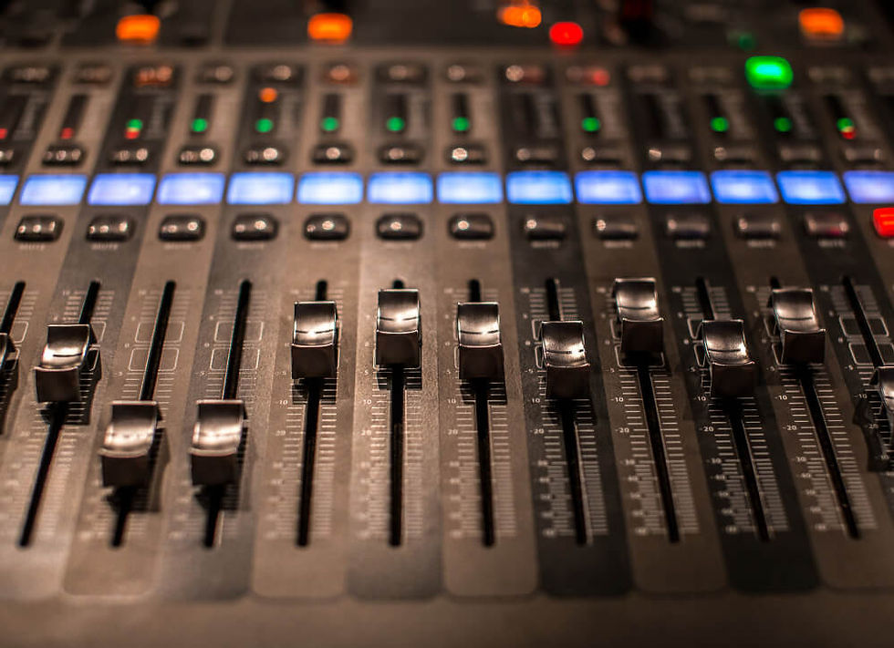A imagem mostra uma mesa de som profissional cheia de controles, botões e sliders. Os botões giratórios (knobs) possuem marcações coloridas em azul, vermelho e branco, usados para ajustar diferentes frequências e efeitos de áudio. Na parte inferior, há faders deslizantes que regulam o volume de cada canal.