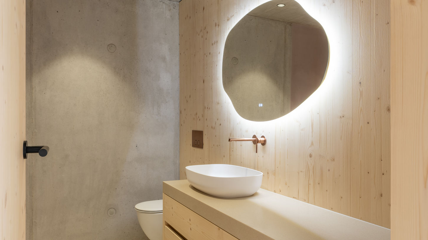 Detail eines runden, beleuchteten Spiegels über einem hellen Holzregal im Bad.