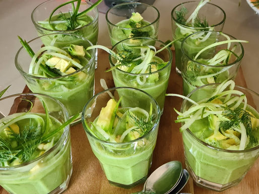 Glasses of green soup garnished with avocado, dill, and cucumber spirals on a wooden board. Several silver spoons nearby.
