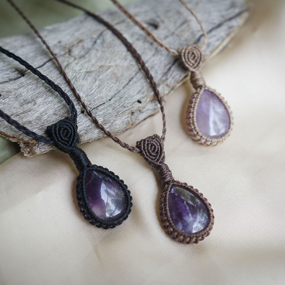 Three teardrop amethyst pendants in black, brown, and tan macramé settings on driftwood, set against a soft beige fabric.