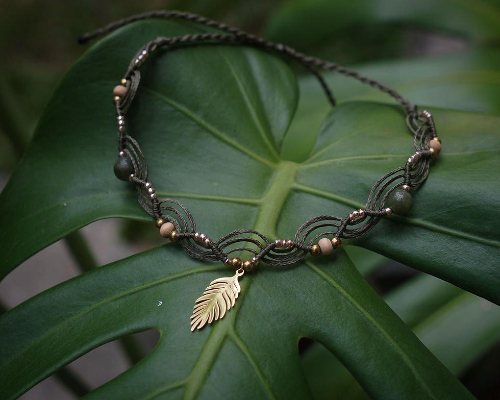 A woven ankle bracelet with Jade beads and a gold leaf pendant rests on a large green leaf, creating a natural and elegant atmosphere.