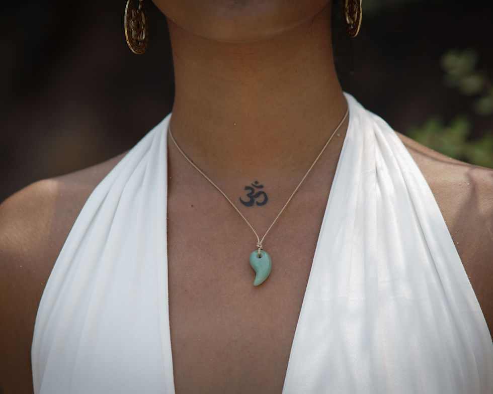 Close-up of a person wearing a white halter top, an 'Om' tattoo on the chest, a magatama pendant necklace, and large earrings. Blurred greenery.