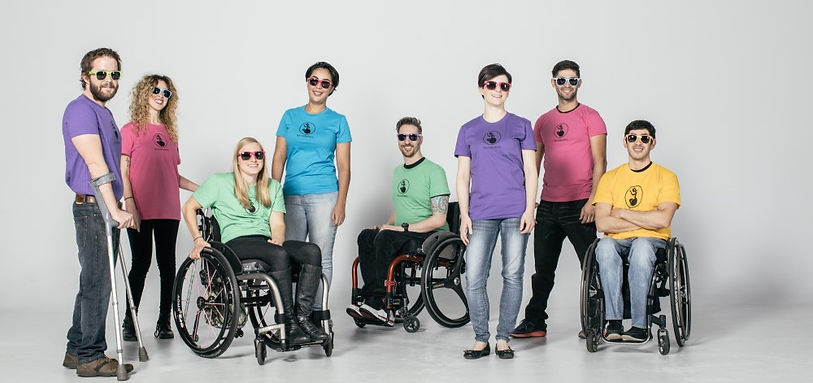 group of people wearing backbones tshirts in different colors ranging from purple, pink, green, blue and yellow. some sit in wheelchairs, others stand, others use a cane or crutches. They all wear sunglasses