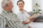 Smiling nurse holding book converses with elderly man, Long Term Care setting.
