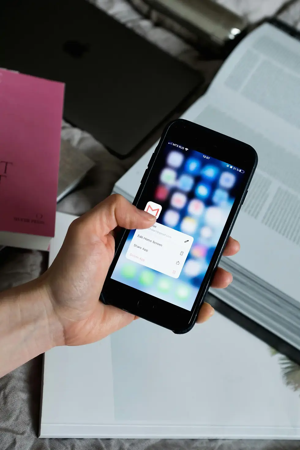 Hand holding a smartphone with blurred app icons and menu options visible. Background has an open book, pink and black items.