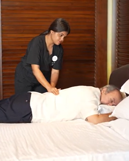 Physiotherapist providing back pain rehab physiotherapy a man lying face down on a white bed. Wooden wall in the background; relaxed atmosphere.