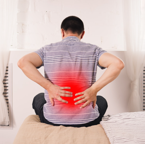 Man sitting on bed holding lower back in pain, highlighted in red. Wearing striped shirt in a bright room with white curtains.