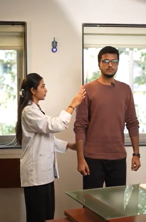 A woman in a lab coat checks a man's posture in a well-lit room. The man wears glasses and a brown sweater, looking focused.
