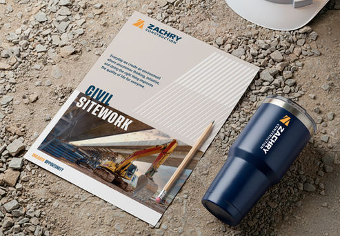 A Zachry brochure on gravel beside a Zachry Construction tumbler, pencil, and hard hat.
