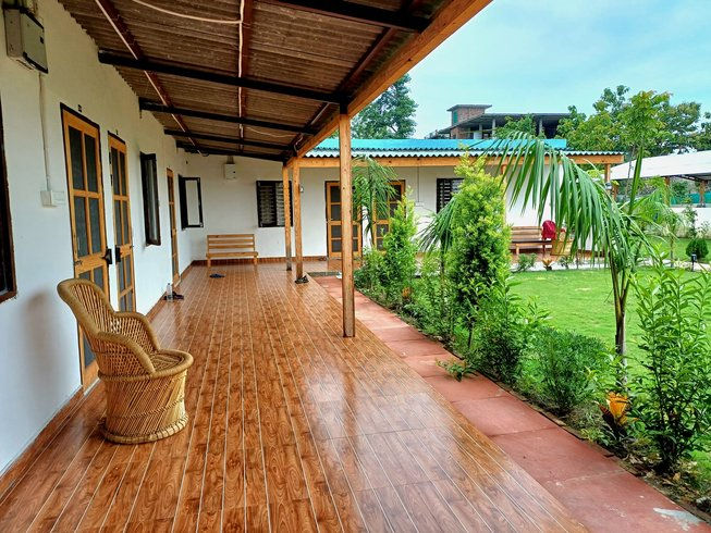 Covered porch with wicker chair and wood-patterned floor, facing a green garden. White buildings in background, sky is slightly overcast. 200 Hour Yoga Alliance Certification Teacher Training Retreat in India