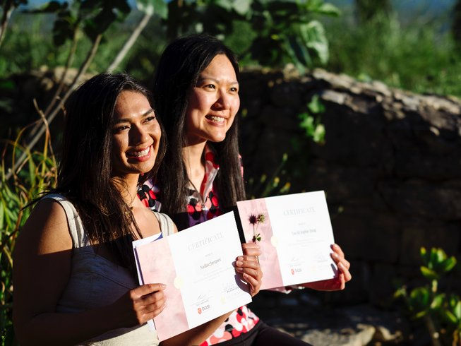 The Best Pilates Teacher Training Retreat 2025. Two women smiling, holding certificates outdoors. Sunlight illuminates them against a backdrop of greenery and a stone wall.