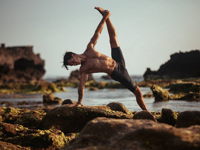 The best summer pilates retreat 2025 in France. Man in shorts performs a yoga pose on rocky sea shore at sunset, stretching arm and leg. Calm and focused expression, warm tones.