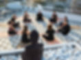 People seated in a circle meditating on a rooftop, near a Buddha statue on white stones. Peaceful atmosphere, patterned mats, and urban backdrop. 200 Hour Yoga Alliance Certification Teacher Training Retreat in Greece
