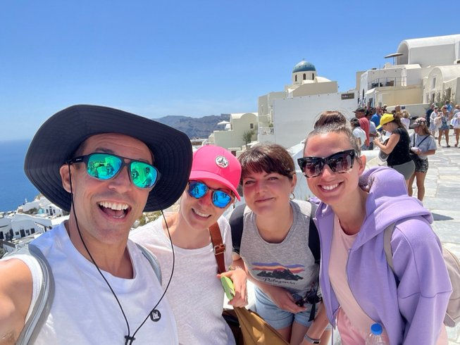 The best summer pilates retreat 2025 in Greece. Four people smile in a sunny, whitewashed town with blue domes, likely Santorini. They're wearing casual clothes and sunglasses. Joyful mood.