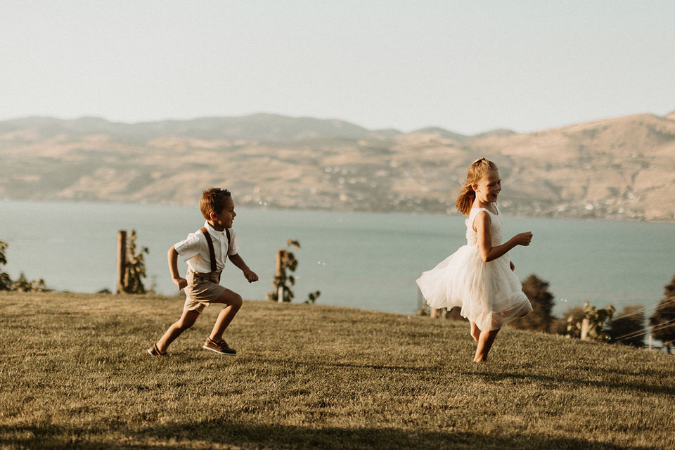 A boy in his tux playing, laughing, and chasing a girl in her flower girl dress with a background of Lake Chelan.