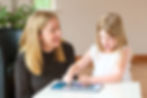 Woman and girl using a speech therapy app on a tablet at a white table. The woman smiles, and the girl points at the screen. Green plant in the background.