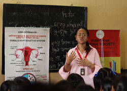 A young female educator from Spriha Society stands in front of a classroom