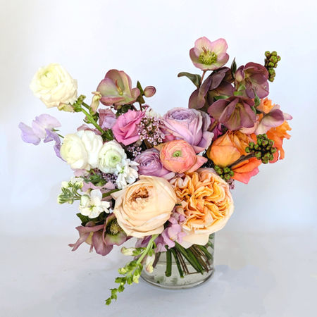 A clear glass vase holds a lush arrangement of pastel and earth-toned flowers, including cream and peach roses, lavender ranunculus, and burgundy hellebores, set againsA clear glass vase holds a lush arrangement of pastel and earth-toned flowers, including lavender and peach roses, cream ranunculus, and burgundy hellebores, set against a plain white background.