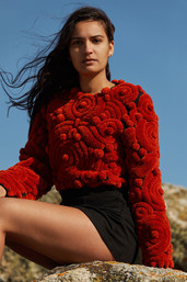 Mannequin femme assise dans les rocher en bretagne avec les cheveux au vent, elle porte un pull brodé rouge s’inspirant des costumes bretons, ainsi qu’une jupe noire.