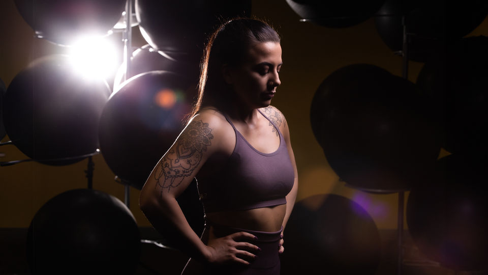 Dramatic fitness branding portrait of a strong, tattooed woman in a sports bra, standing confidently in a dimly lit gym. Backlighting creates a powerful silhouette with lens flares adding intensity. A bold image perfect for personal branding, fitness marketing, and athlete promotion. Photo by North Star Images.
