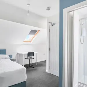 Property Photography North East is a folio of North Star Images
Specialising in Serviced accommodation photography, Interior Photographer HMO Photography Redcar
Bright loft bedroom with ensuite shower room, featuring a double bed with blue upholstered headboard, white bedding, skylight roof window, small workspace with desk and chair, grey carpet, and modern glass shower enclosure clean, modern serviced accommodation interior photographed by property Photography North East a folio of North Star Images.