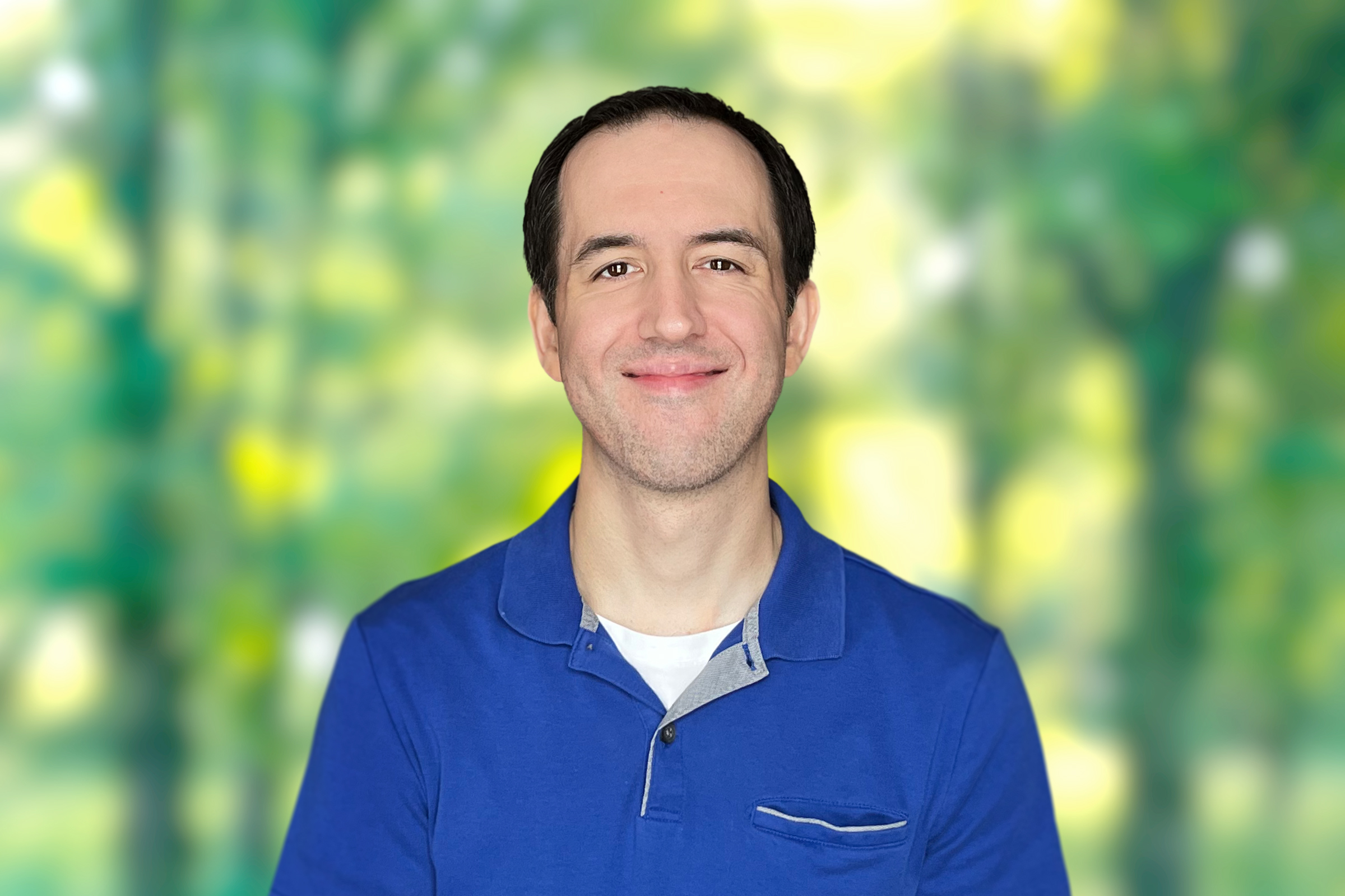 Smiling man in blue shirt standing in front of a blurred background.