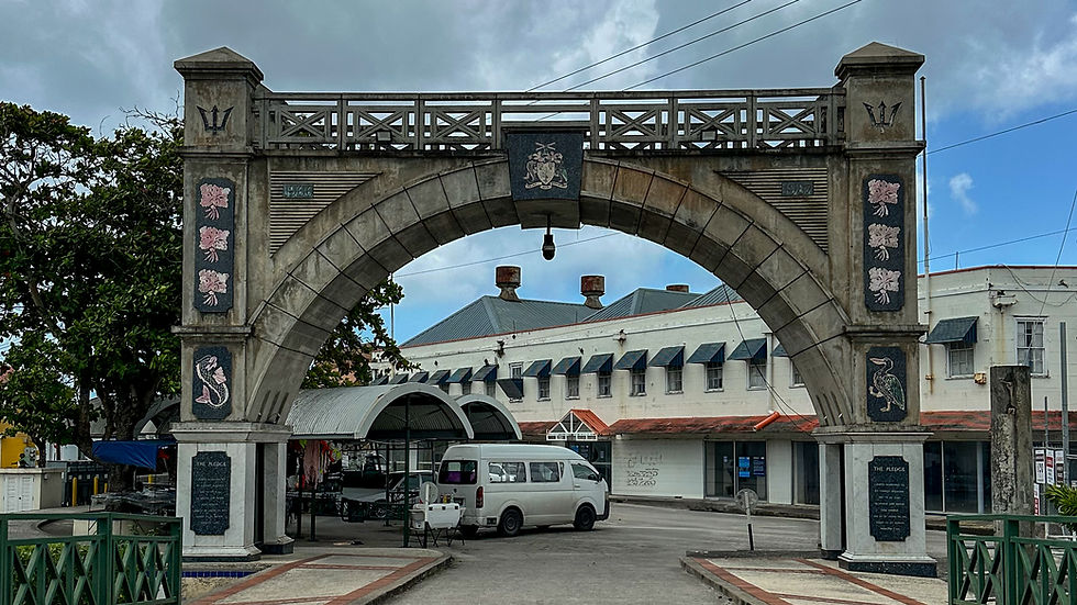 Stone archway with intricate designs, flanked by trees, over a street with a white van parked. Background features a building with blue awnings.