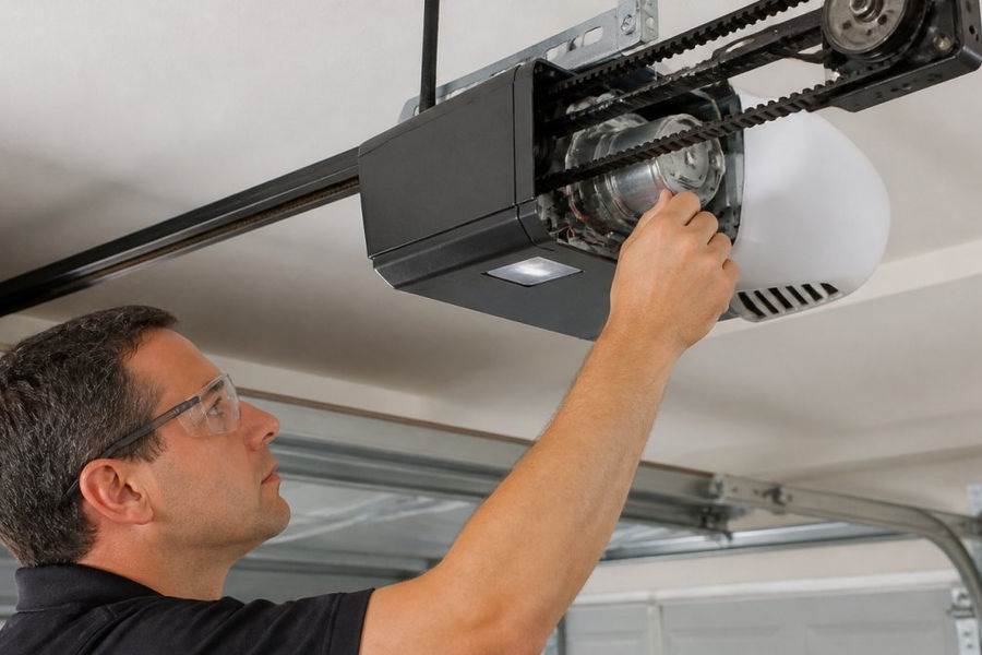 Professional technician examining a residential garage door opener motor mounted on the ceiling, showing the belt drive system, pulleys, and mechanical components in a modern garage