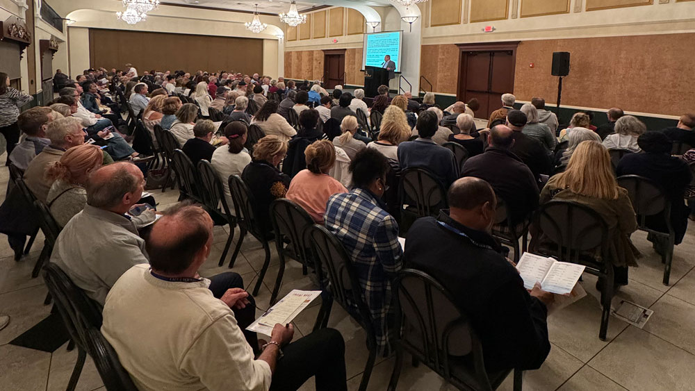 A large audience sits in a spacious hall, attentively listening to a speaker at a podium. A blue presentation screen is displayed.