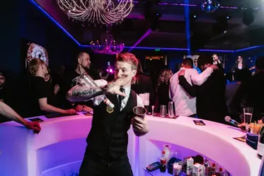 Las Vegas flair bartenders performing behind a curved illuminated bar during a high-energy nightlife event, confidently working bottles, shakers, and glassware while engaging surrounding guests. The image highlights professional Las Vegas flair bartenders trained to balance entertainment and service, delivering visually dynamic bar shows without slowing drink production. Ideal for corporate events, private parties, brand activations, and upscale nightlife experiences where flair bartending adds movement, rhythm, and guest interaction to the atmosphere.