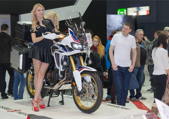Los Angeles trade show staff member presenting a featured motorcycle at a major moto exhibition in Los Angeles, standing beside a branded motorcycle display and engaging attendees on the show floor. The image highlights professional trade show staff supporting brand visibility, product presentation, and guest interaction during large-scale expos and conventions. This Los Angeles trade show staff professional assists exhibitors by managing display areas, answering visitor questions, and maintaining polished brand representation throughout trade shows and industry events in Los Angeles 2026.