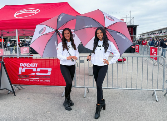 Promo models representing a motorsports brand during a San Francisco Bay Area automotive showcase, supporting attendee engagement, sponsor visibility, and guest interaction at a large-scale outdoor racing event near the waterfront.