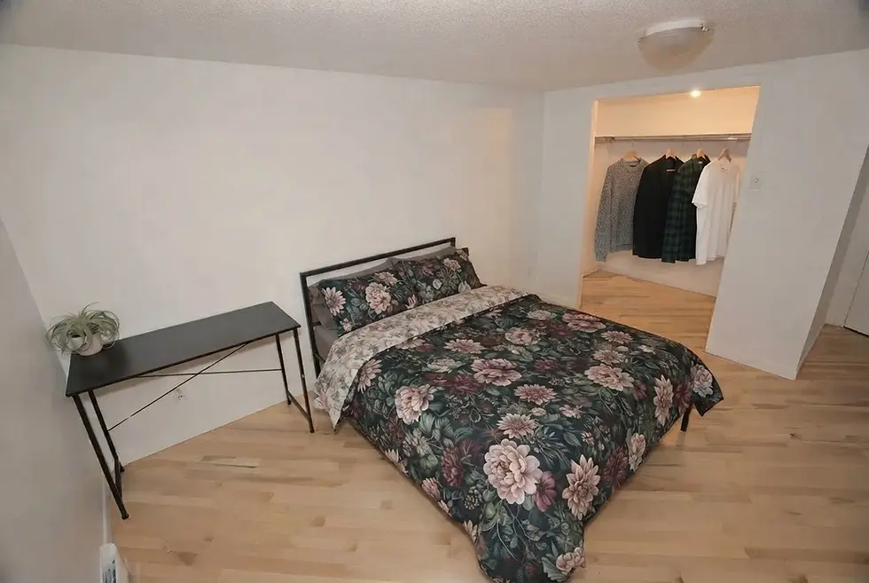 Chambre meublée à Jonquière avec lit double, literie fleurie, table d'appoint et rangement ouvert.