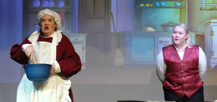 Mrs. Brill (Kristi Hibschman) stirs something in a blue bowl while Robertson Ay looks on and talks to her from behind. They are standing in the kitchen.
