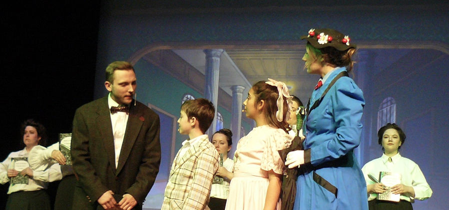 Northbrook (John Roniger) talks with Michael and Jane Banks in the bank while Mary looks on, with the bankers in the background.