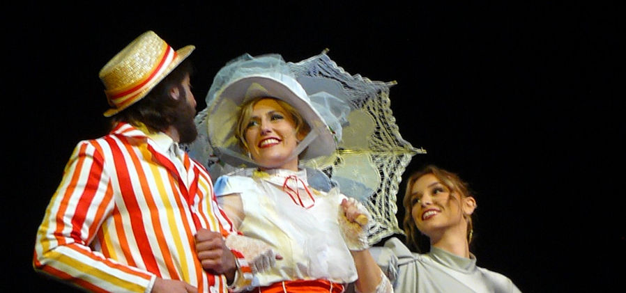 Bert (Andrew Ainsworth), Mary (Anna O'Renick) and Neleus (Audrey Phillips) dance arm in arm. Mary wears her classic white dress with a red sash, while Bert is dressed in a red and yellow striped jacket.