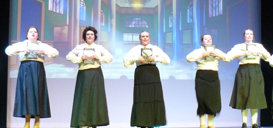 Miss Smythe (Marie Hall) and the bank staff stand in a line, holding writing tablets, dressed in black skirts and white blouses.