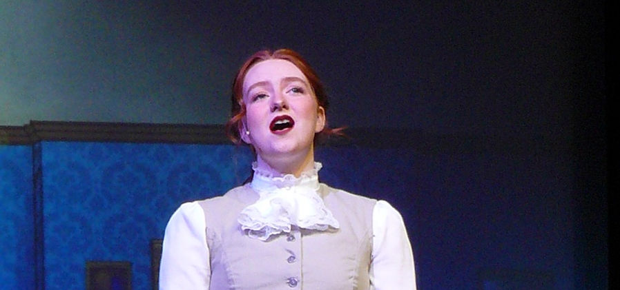 Winifred Banks (Emily Turner) stands in her living room, singing while wearing her tan Victorian dress.