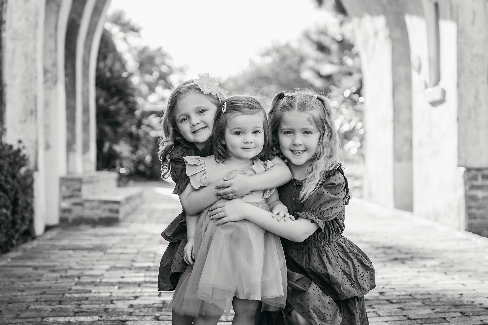 A black and white photo of two little girls leaning over on each side of their two year old sister to give her a hug as they all smile at the camera, standing on a brick paved walkway at Casa Feliz in Winter Park, FL