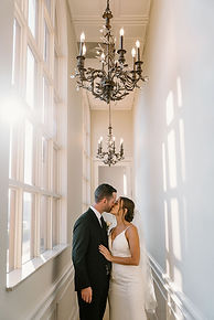 A bride and groom kiss as they stand in the middle of a sunlit halway with a chandellier above them at Pier 22 in Bradenton, Florida