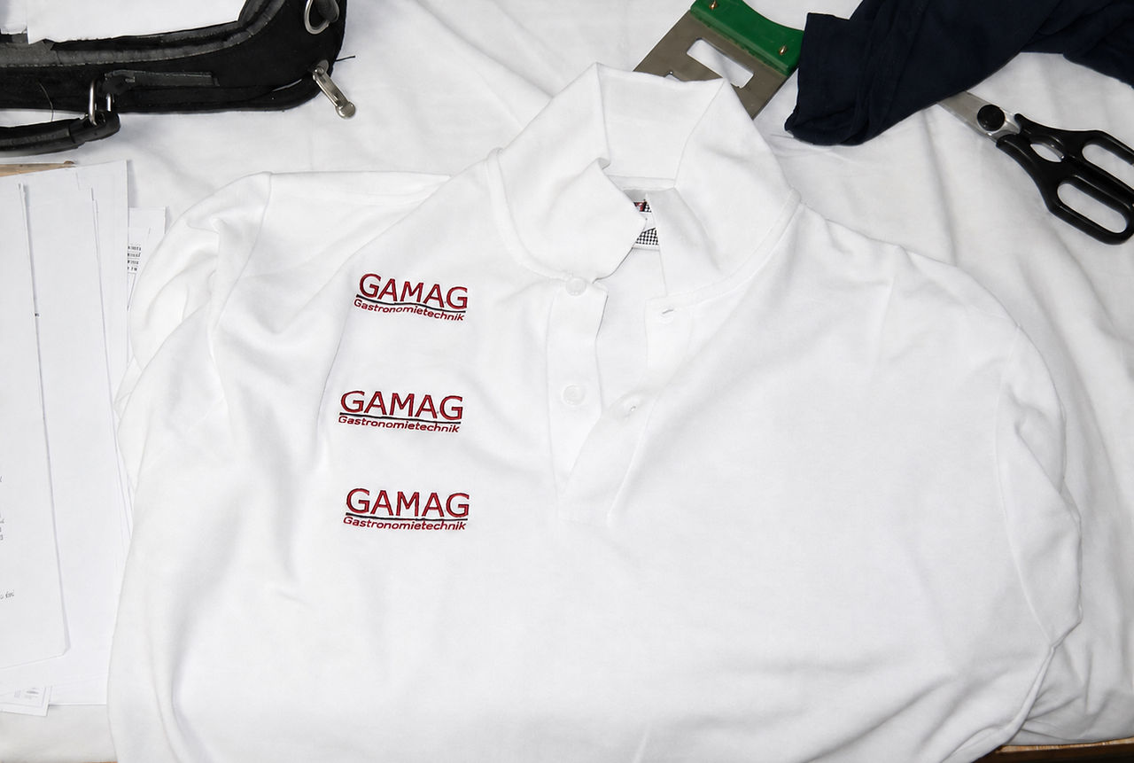 Mehrere weiße Poloshirts mit aufgestickten Firmenlogos liegen auf einem Arbeitstisch.